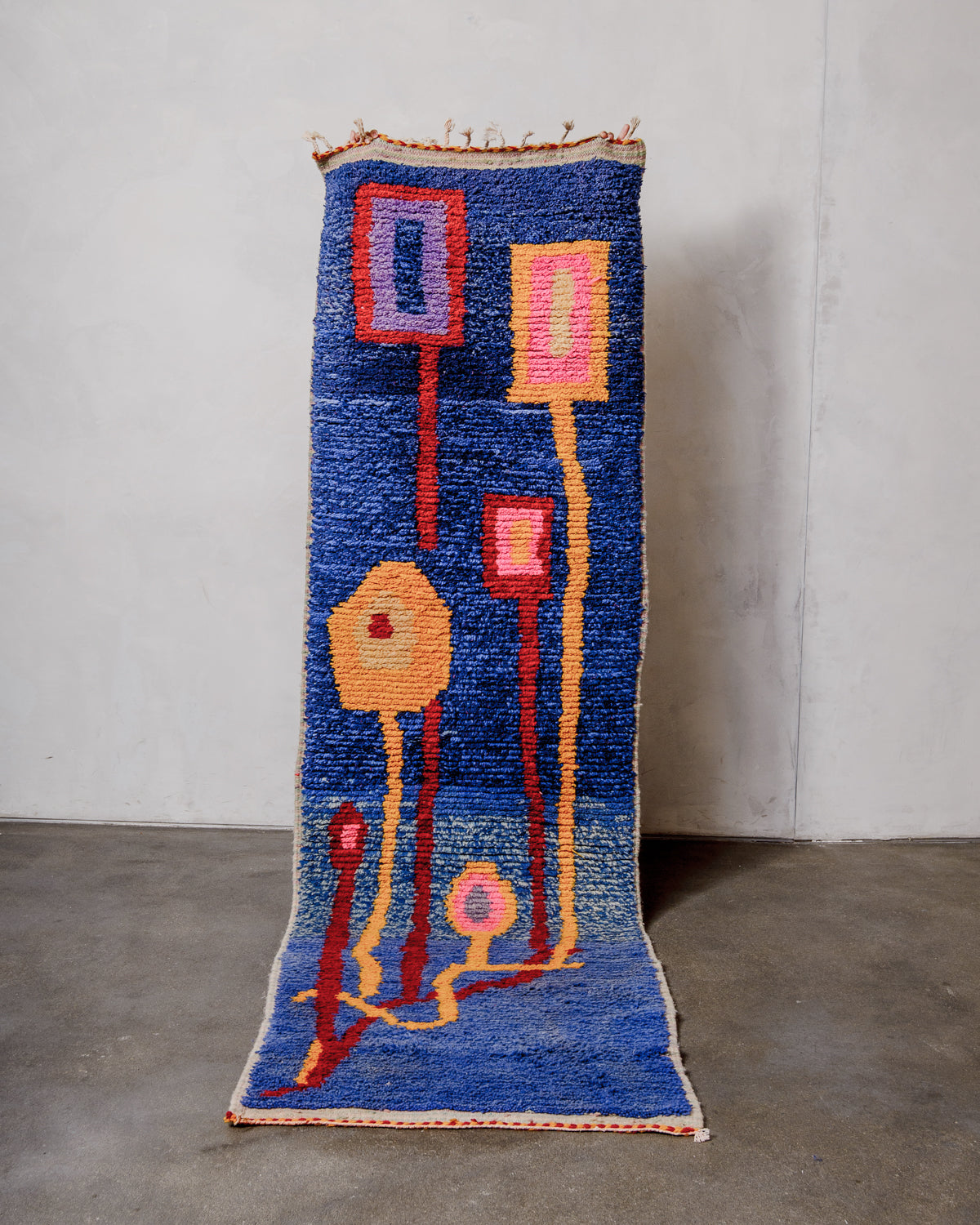 Berberlin vintage runner rug made of brightly coloured wool, being held up in front of a white wall and concrete floor.